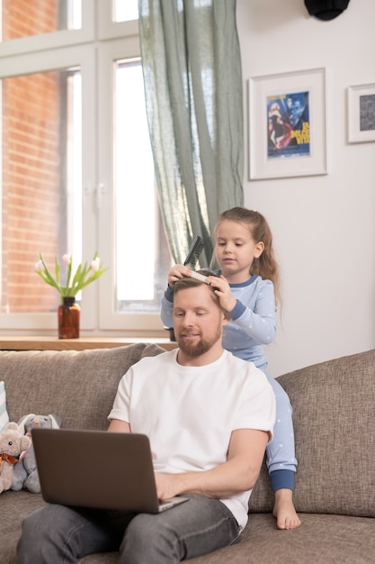 Bambina sveglia che spazzola i capelli di suo padre con il computer portatile che lavora a distanza mentre entrambi seduti sul divano morbido dalla finestra nel soggiorno