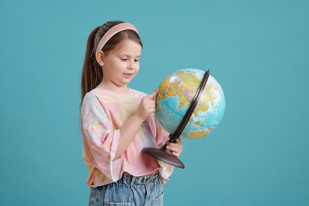 Bambina sveglia che tiene il globo nelle sue mani e lo esamina in piedi sullo sfondo blu