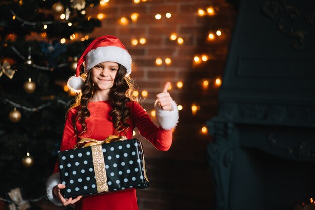 Bambina sveglia che tiene un regalo vicino all'albero di Natale in un costume rosso di babbo natale