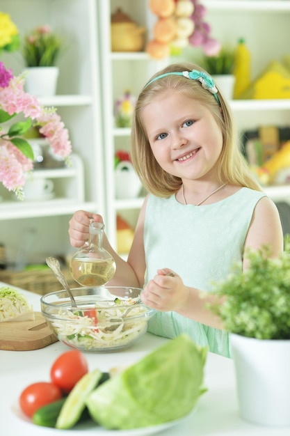 Bambina sveglia che versa olio in insalata fresca sul tavolo della cucina a casa