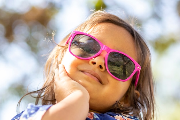 Bambina sveglia con gli occhiali da sole rosa e le mani sul suo viso.