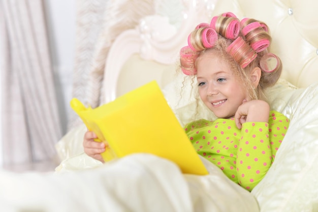 Bambina sveglia con i bigodini rosa che si trovano in cattivo e libro di lettura