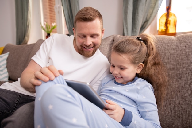 Bambina sveglia con il touchpad e suo padre che guardano attraverso film o cartoni animati online mentre trascorrono del tempo a casa in quarantena