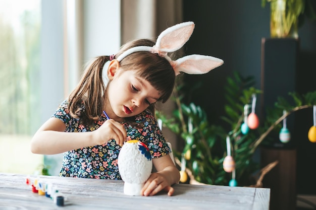 Bambina sveglia con le orecchie del coniglietto che dipinge l'uovo di Pasqua con il pennello