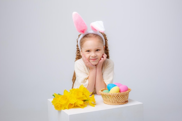 Bambina sveglia con orecchie da coniglio su sfondo bianco con un cesto di uova di Pasqua e fiori