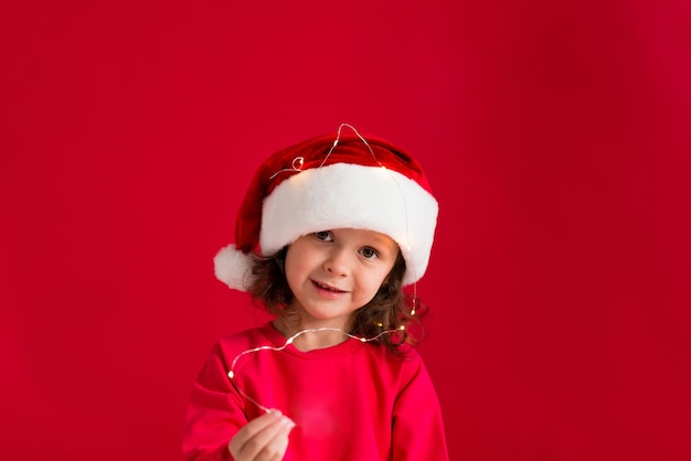 Bambina sveglia con un cappello rosso di Natale e un vestito rosso su sfondo rosso. Buon Natale