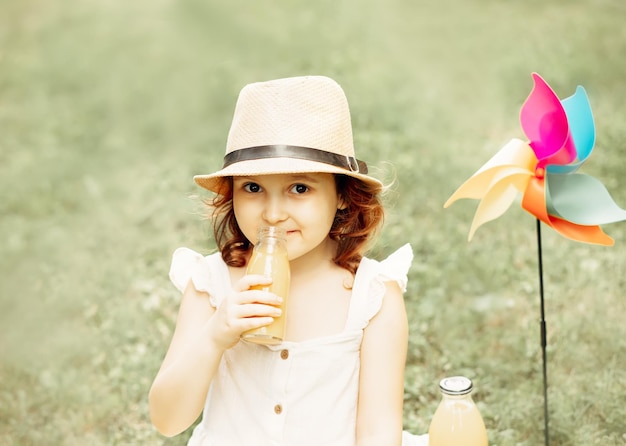 Bambina sveglia con un cappello seduto su una coperta su un prato del parco che tiene una bottiglia e beve succo d'arancia Picnic sulla natura