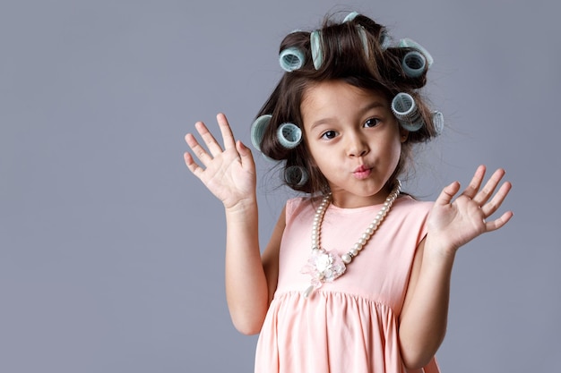 Bambina sveglia divertente in abito rosa e bigodini in posa su sfondo grigio. Emozioni umane ed espressione facciale