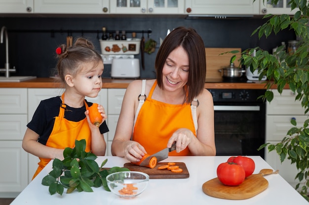 Bambina sveglia e bella madre in grembiule arancione stanno cucinando, tagliando, tritando le verdure, sorridendo, divertendosi al fondo della cucina, spazio della copia