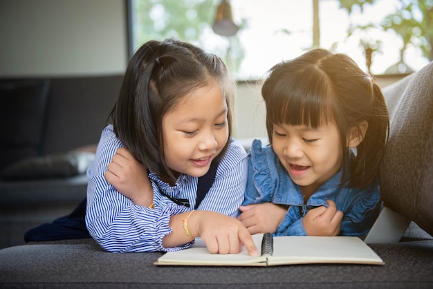 Bambina sveglia in abbigliamento casual che legge un libro e che sorride mentre trovandosi su un sofà nella stanza.