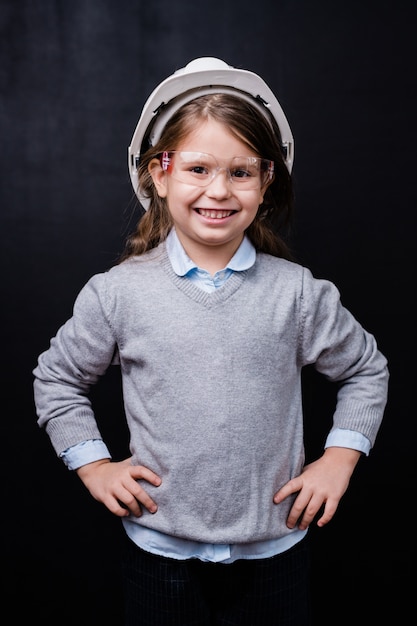 Bambina sveglia in elmetto protettivo e occhiali protettivi mantenendo le mani sulla vita in isolamento