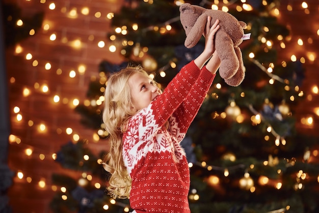 Bambina sveglia in maglione festivo rosso con l'orsacchiotto all'interno che celebra il nuovo anno e le feste di natale.