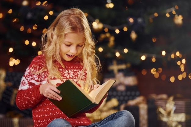 Bambina sveglia in maglione rosso festivo libro di lettura al chiuso durante le vacanze di natale.