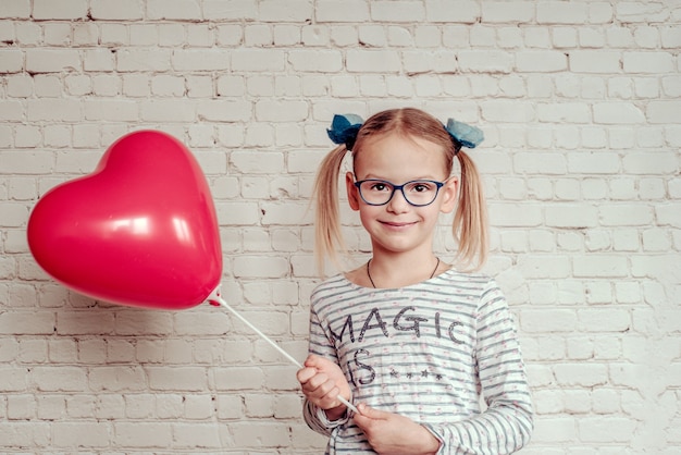 Bambina sveglia in occhiali con palloncino a forma di cuore rosso su sfondo bianco muro di mattoni, sfondo di San Valentino