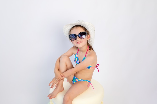 Bambina sveglia in occhiali da sole neri e bikini blu è seduta sul pavimento