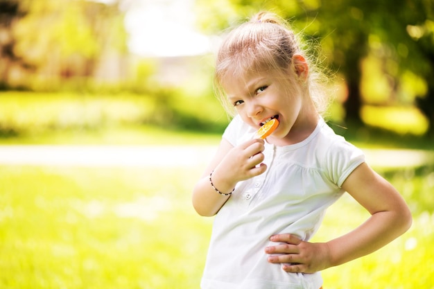 Bambina sveglia in piedi sull'erba verde nel parco e tenendo in mano lecca-lecca arancione.