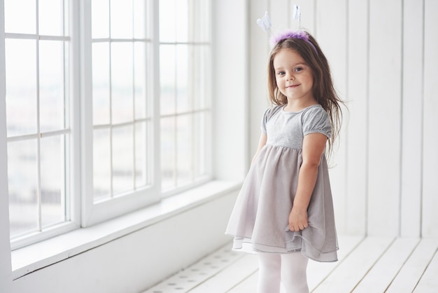 Bambina sveglia in vestito che sta nella stanza bianca vicino alle finestre.