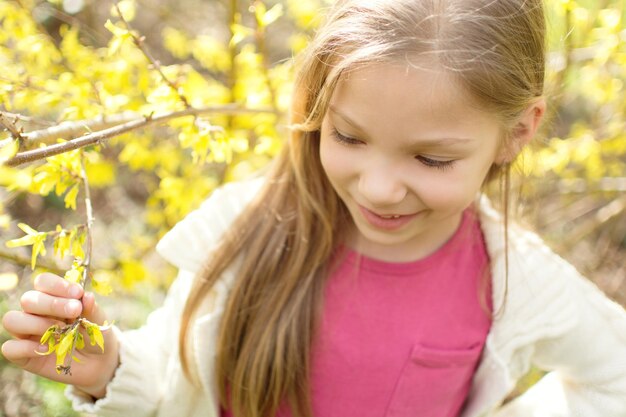 Bambina sveglia sorridente in piedi accanto a un albero di fiori nel parco e pensare.