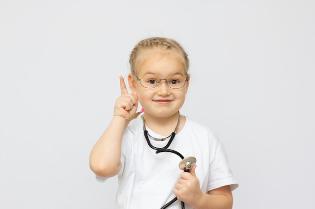 Bambina sveglia vestita come un medico che guarda l'obbiettivo con un sorriso allegro dito verso l'alto