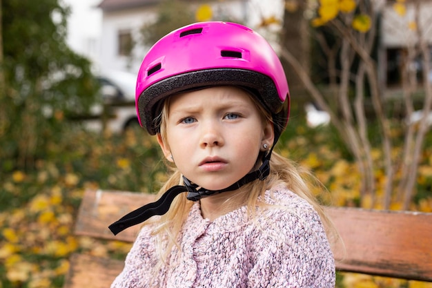 Bambina triste in un casco rosa seduto su una panchina del parco