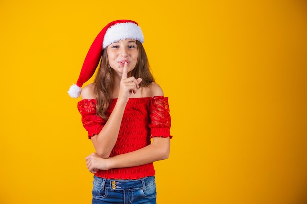 Bambina vestita per natale facendo segno di silenzio su sfondo giallo.