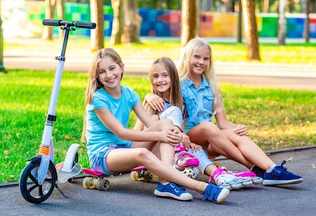 Bambine abbastanza sorridenti che si siedono vicino all'erba nel parco serale di sole con scooter e pattini a rotelle