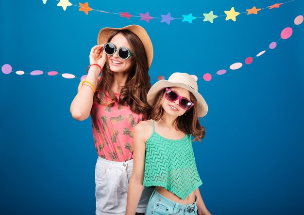 Bambine carine allegre in occhiali da sole in piedi e in posa su sfondo blu
