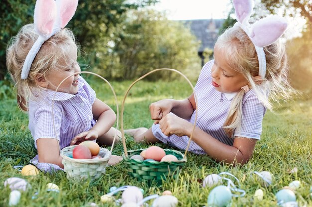 Bambine carine in divertenti orecchie da coniglio giocano in un gioco raccogliendo uova di Pasqua in un cesto di vimini su un'erba su sfondo verde sfocato Concetto di vacanze primaverili