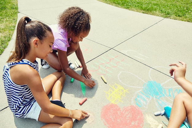 Bambine che disegnano a terra con il gesso a scuola per l'apprendimento, lo sviluppo o la creatività Arte di gruppo e bambini al sole sul marciapiede o scrivendo con linee e modelli di fiori al parco