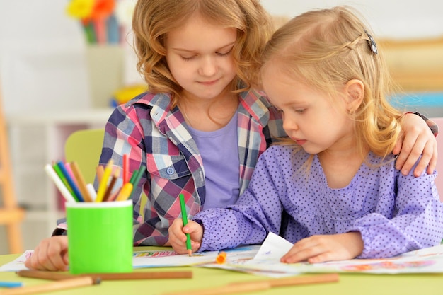 Bambine sveglie che si siedono al tavolo e che disegnano con le matite nella loro stanza