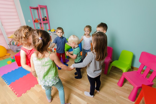 Bambini allegri in età prescolare in piedi in cerchio che si tengono per mano Giochi per insegnare la cooperazione e il team building Bambini adorabili in una stanza con mobili colorati