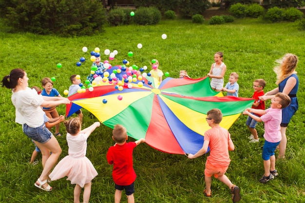 Bambini attivi che sventolano un grande paracadute multicolor per lanciare le palle Giochi di gruppo per rafforzare la relazione tra i bambini Parco estivo con erba verde Sbarazzarsi di cose inutili concept