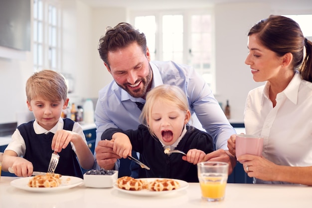 Bambini che indossano l'uniforme scolastica in cucina che mangiano cialde per la colazione mentre i genitori si preparano per il lavoro