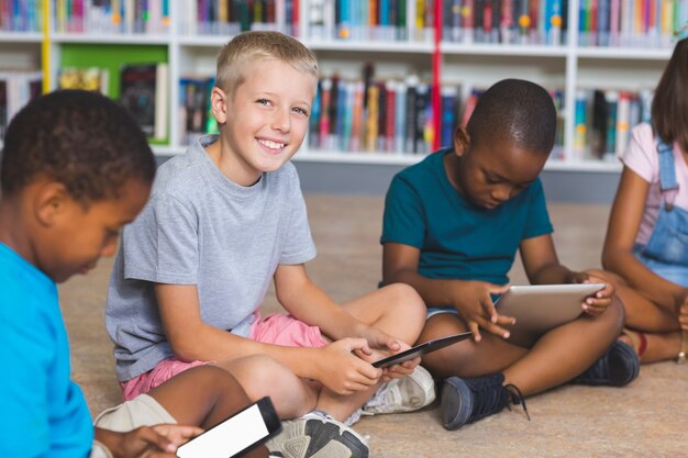 Bambini della scuola che si siedono sul pavimento facendo uso della compressa digitale nella biblioteca