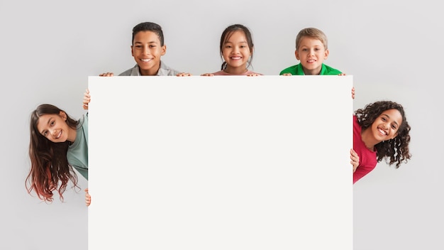 Bambini diversi felici che si nascondono dietro uno sfondo grigio vuoto del bordo bianco