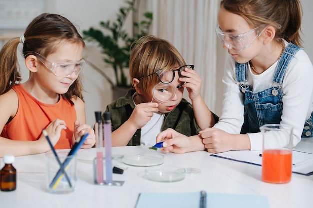 Bambini divertenti che fanno un progetto di scienze domestiche, guardando una foglia con una pinzetta. Ragazzo divertente che guarda tramite la lente d'ingrandimento. Tutti dietro il tavolo, con gli occhiali. Vetreria chimica e liquidi colorati sul tavolo.