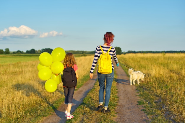 Bambini due ragazze che camminano lungo la strada campestre con cane e palloncino, vista posteriore