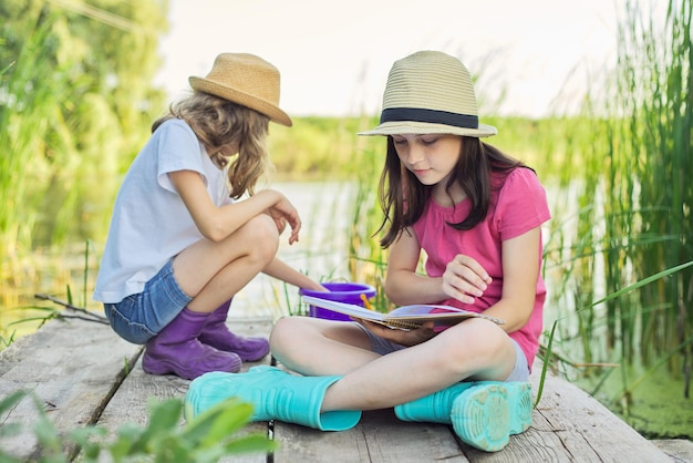 Bambini due ragazze che riposano giocando a leggere il loro taccuino in natura. Bambini seduti sul molo di legno del lago, sfondo del paesaggio dell'acqua del tramonto estivo, stile country