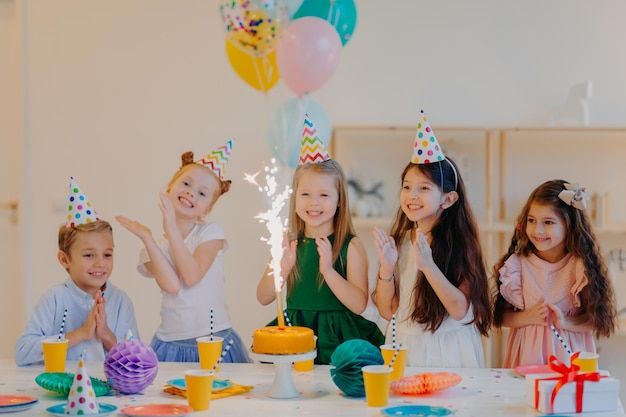 Bambini e concetto di vacanza Sono contento che cinque amici guardino con gioia sulla torta con scintillio festeggiano il compleanno indossano cappelli a cono e tengono palloncini d'aria hanno espressioni felici