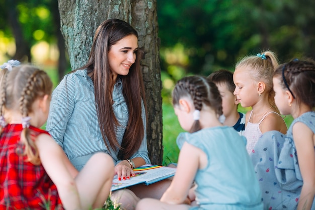 Bambini e istruzione, giovane donna al lavoro come educatore, leggendo un libro per ragazzi e ragazze nel parco