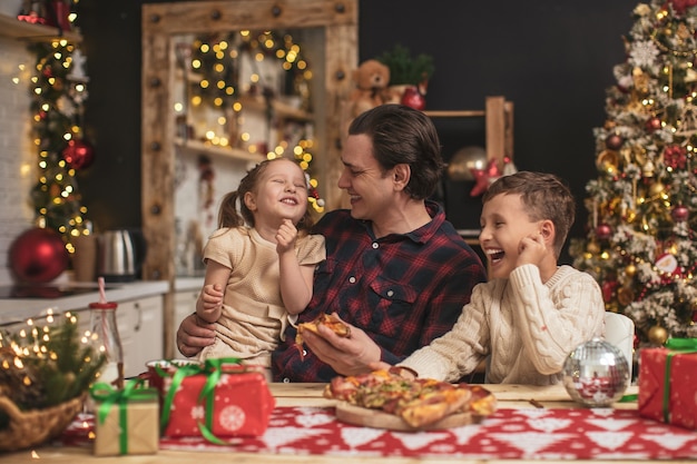 Bambini felici con papà che mangia pizza in cucina decorata per Natale