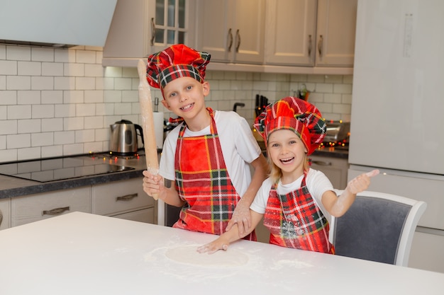 Bambini felici cucinano i biscotti di Natale a casa in cucina in un costume da chef, un piccolo ragazzo e una ragazza biondi