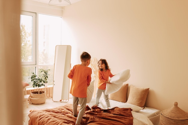 Bambini felici e divertenti fratello e sorella in pigiama che giocano a combattere con i cuscini in una camera da letto accogliente a casa