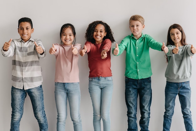 Bambini multietnici felici che gesticolano i pollici in su in posa in piedi all'interno