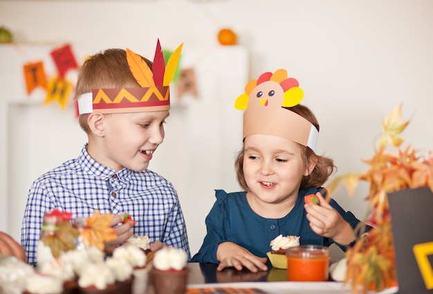 Bambini seduti sul tavolo festivo e celebrano il giorno del Ringraziamento. Bambini in cappello di tacchino di carta e cappelli da pellegrino che mangiano cupcakes e bevono latte.