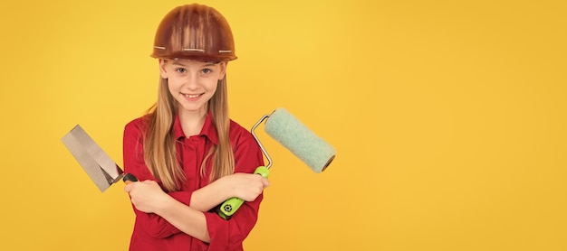 Bambino adolescente positivo in elmetto da costruttore con rullo di vernice e spatola sulla parete gialla Generatore di bambini nel design del poster orizzontale del casco Spazio per la copia dell'intestazione banner