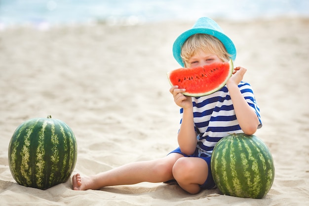 Bambino adorabile alla riva di mare che mangia anguria succosa. Bambino allegro in estate sulla spiaggia. Ragazzino sveglio all'aperto