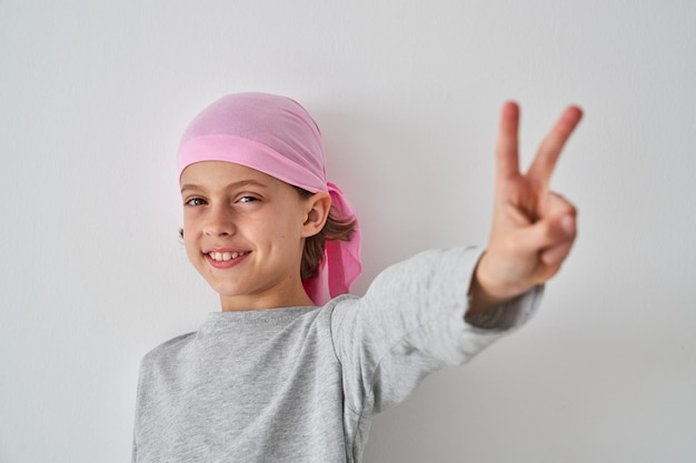 Bambino adorabile allegro in bandana rosa che mostra il segno V e sorridente mentre sta in piedi su sfondo bianco e guarda la macchina fotografica