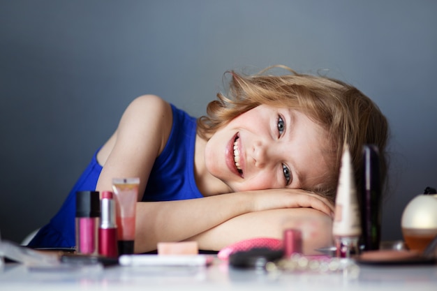 Bambino affascinante, piccola bellezza, ragazza carina di 7-8 anni con bellissimi riccioli biondi, trucco, trucco della mamma al tavolo, muro grigio