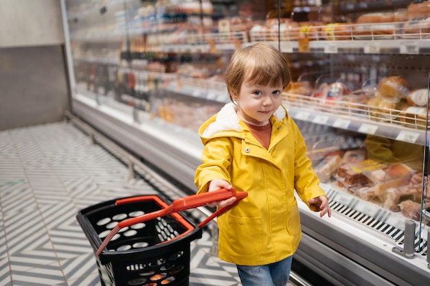 Bambino al mercato con un carrello della spesa che indossa una giacca gialla e jeans